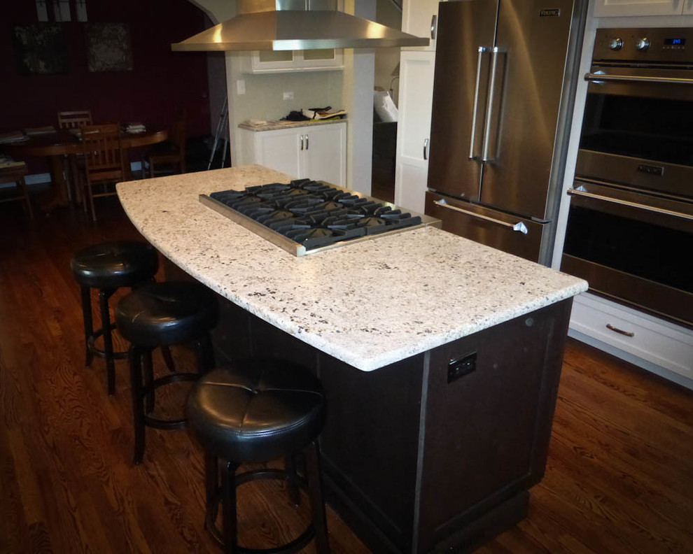 Klassische Küche in L-Form mit Schrankfronten im Shaker-Stil, weißen Schränken, Granit-Arbeitsplatte, Küchengeräten aus Edelstahl, braunem Holzboden und Kücheninsel in Denver