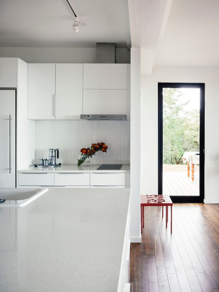 На фото: кухня в стиле модернизм с плоскими фасадами, белыми фасадами, столешницей из переработанного стекла, белым фартуком, фартуком из плитки кабанчик, паркетным полом среднего тона и островом