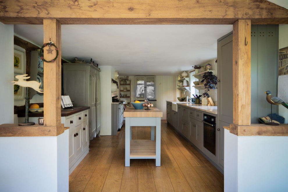 This is an example of a medium sized farmhouse galley kitchen/diner in West Midlands with a belfast sink, shaker cabinets, grey cabinets, black appliances, medium hardwood flooring, an island, brown floors and beige worktops.