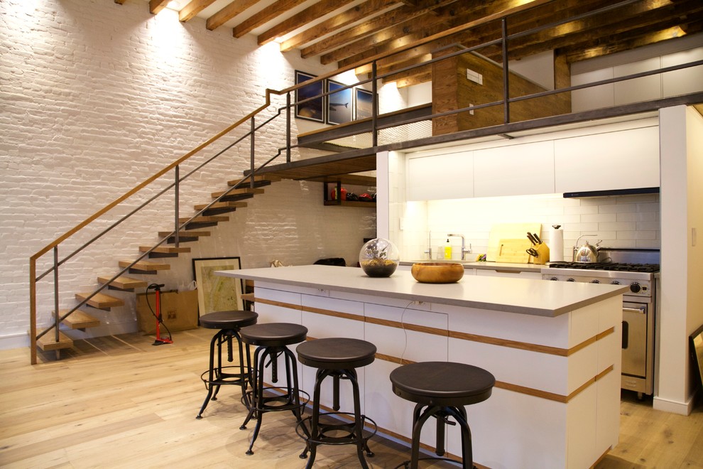 Example of a mid-sized urban single-wall light wood floor open concept kitchen design in New York with flat-panel cabinets, an island, a drop-in sink, white cabinets, white backsplash, stainless steel appliances and subway tile backsplash