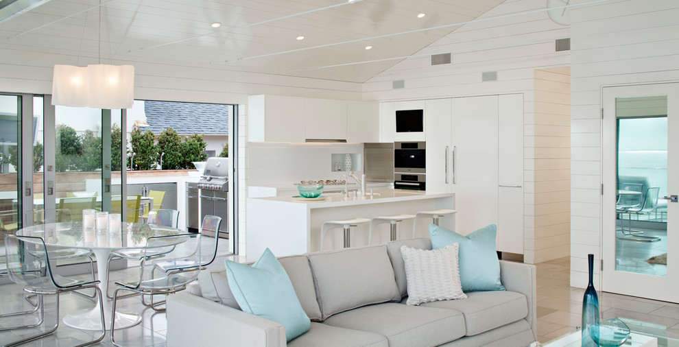 На фото: угловая кухня-гостиная в морском стиле с плоскими фасадами, белыми фасадами, белым фартуком и техникой под мебельный фасад