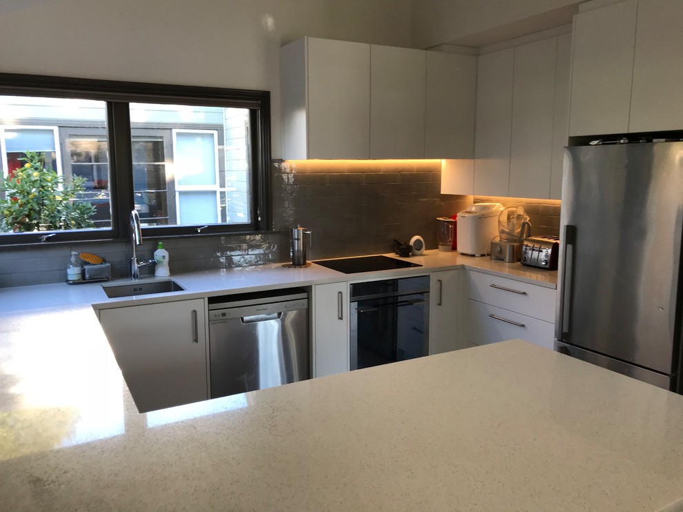 Example of a mid-sized trendy u-shaped vinyl floor and brown floor open concept kitchen design in Christchurch with a single-bowl sink, flat-panel cabinets, white cabinets, quartz countertops, gray backsplash, subway tile backsplash, stainless steel appliances and white countertops
