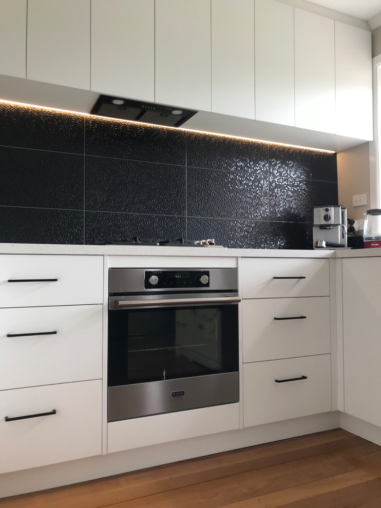 Medium sized contemporary u-shaped enclosed kitchen in Christchurch with a double-bowl sink, flat-panel cabinets, white cabinets, laminate countertops, black splashback, porcelain splashback, stainless steel appliances, medium hardwood flooring, no island and brown floors.
