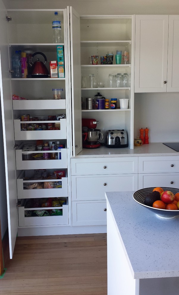 Photo of a medium sized traditional galley open plan kitchen in Christchurch with a double-bowl sink, recessed-panel cabinets, white cabinets, engineered stone countertops, white splashback, glass sheet splashback, white appliances, medium hardwood flooring, an island, brown floors and white worktops.