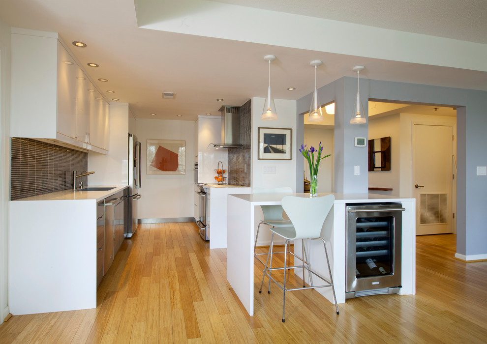 На фото: параллельная кухня-гостиная среднего размера в современном стиле с одинарной мойкой, плоскими фасадами, белыми фасадами, серым фартуком, техникой из нержавеющей стали, паркетным полом среднего тона и островом