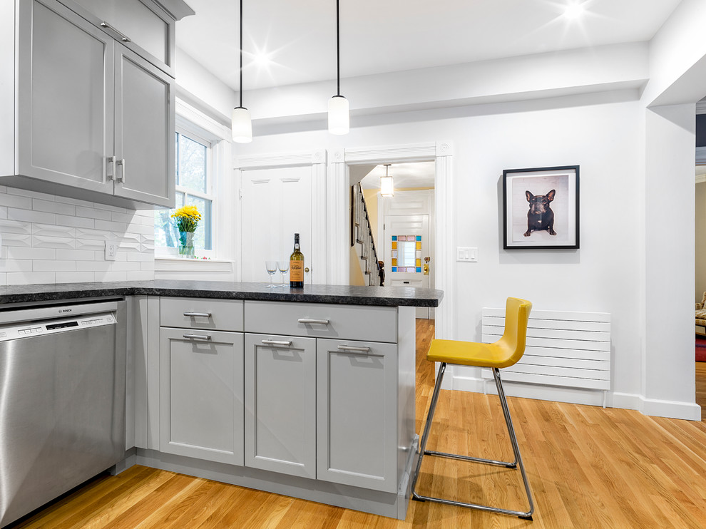 Example of a transitional l-shaped medium tone wood floor and brown floor kitchen design in Boston with shaker cabinets, gray cabinets, white backsplash, subway tile backsplash, stainless steel appliances, a peninsula, black countertops and a farmhouse sink