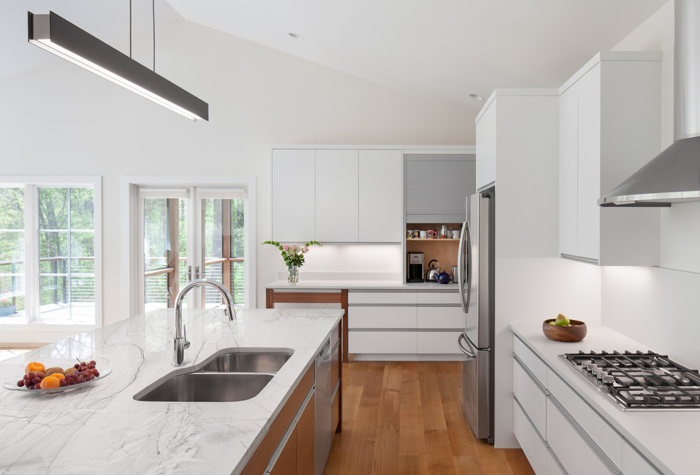 Design ideas for a large contemporary galley open plan kitchen in Raleigh with a double-bowl sink, flat-panel cabinets, white cabinets, quartz worktops, white splashback, stainless steel appliances, medium hardwood flooring, an island and white worktops.
