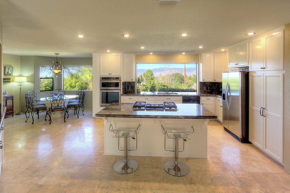 Example of a large trendy l-shaped travertine floor eat-in kitchen design in Los Angeles with an undermount sink, shaker cabinets, white cabinets, granite countertops, black backsplash, stone slab backsplash, stainless steel appliances and an island