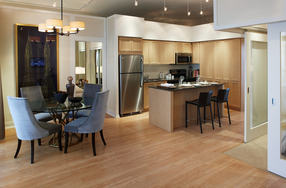 Пример оригинального дизайна: угловая кухня-гостиная в современном стиле с фасадами с утопленной филенкой, светлыми деревянными фасадами, белым фартуком и техникой из нержавеющей стали