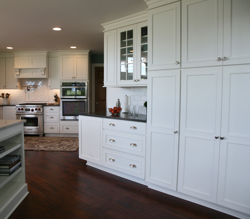 This is an example of a large classic kitchen in Milwaukee with recessed-panel cabinets, white cabinets, engineered stone countertops, white splashback, porcelain splashback, stainless steel appliances, dark hardwood flooring and an island.