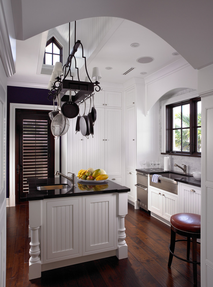 Elegant dark wood floor enclosed kitchen photo in Miami with a farmhouse sink, white cabinets, granite countertops, stone slab backsplash and an island