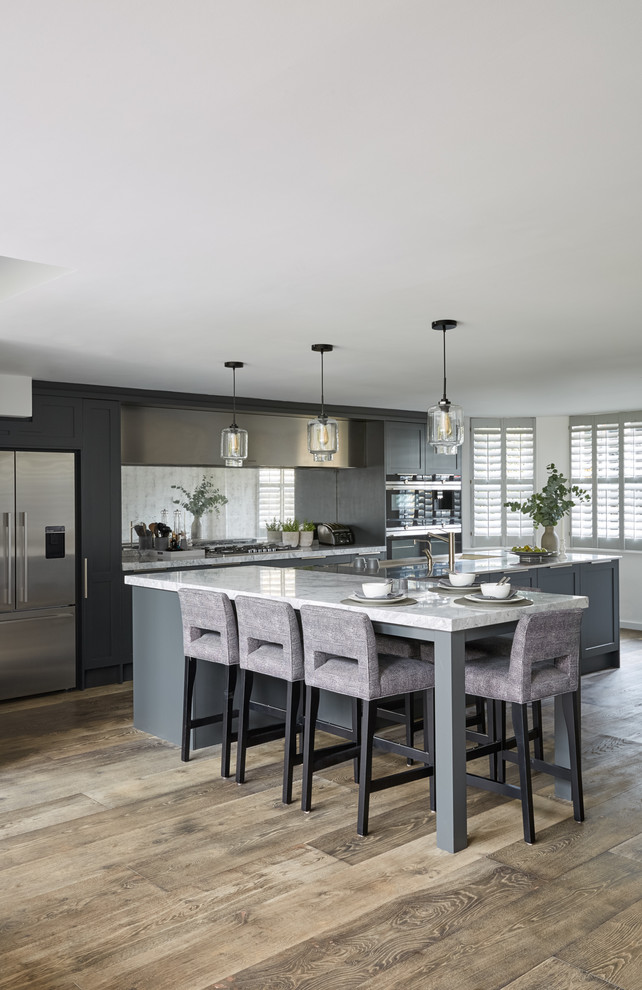 This is an example of a large contemporary galley open plan kitchen in London with an integrated sink, recessed-panel cabinets, grey cabinets, marble worktops, metallic splashback, mirror splashback, stainless steel appliances, dark hardwood flooring, a breakfast bar, brown floors and white worktops.