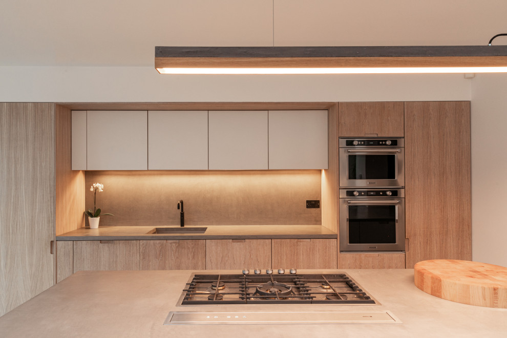 Example of a mid-sized trendy galley open concept kitchen design in London with flat-panel cabinets, light wood cabinets, concrete countertops, an island, gray countertops, an undermount sink, beige backsplash and paneled appliances