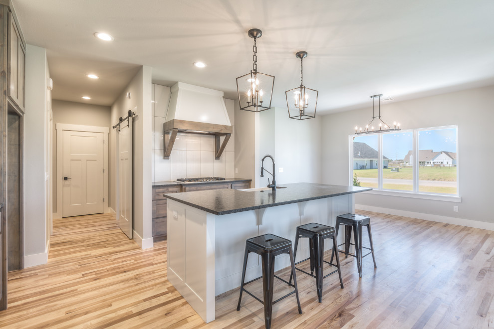 Example of a mid-sized cottage l-shaped medium tone wood floor and brown floor open concept kitchen design in Other with a farmhouse sink, shaker cabinets, medium tone wood cabinets, white backsplash, porcelain backsplash, stainless steel appliances, an island and black countertops