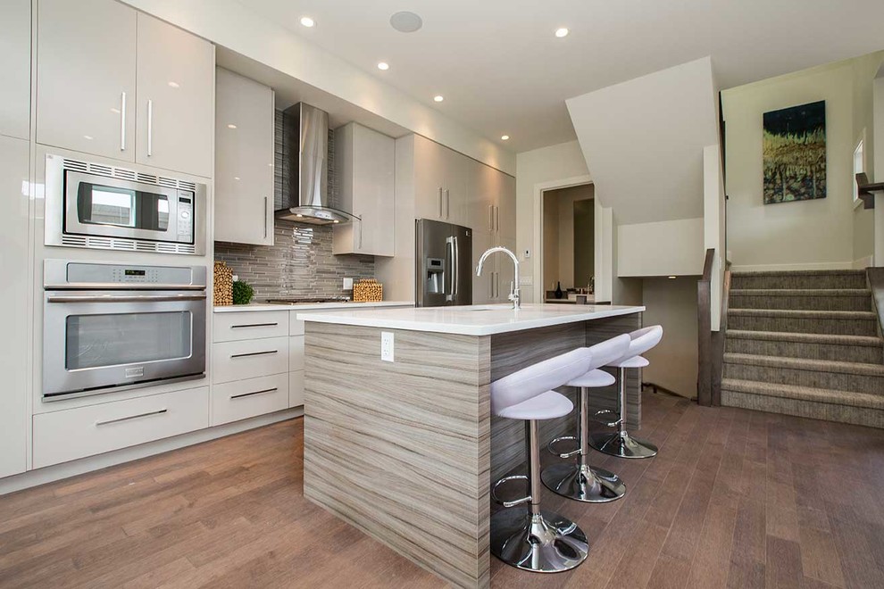 Photo of a medium sized contemporary single-wall open plan kitchen in Calgary with flat-panel cabinets, an island, beige cabinets, composite countertops, beige splashback, matchstick tiled splashback, stainless steel appliances, medium hardwood flooring, a double-bowl sink and brown floors.