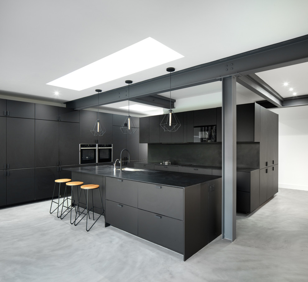 Photo of an urban l-shaped kitchen in London with a submerged sink, flat-panel cabinets, black cabinets, black splashback, black appliances, concrete flooring, an island, grey floors and black worktops.