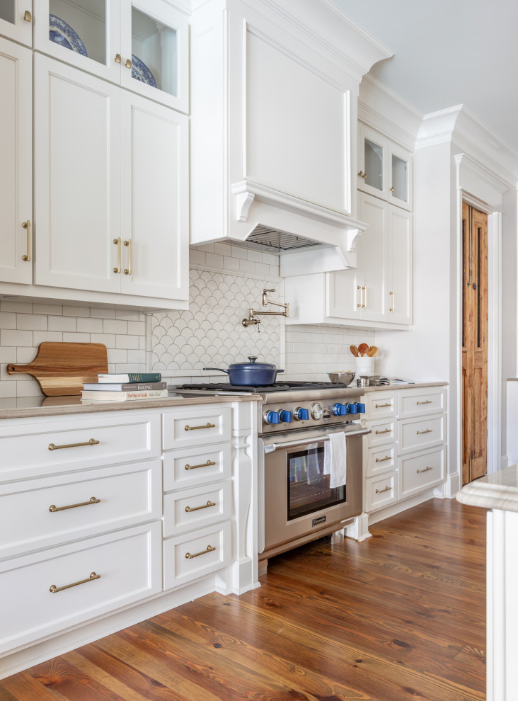Design ideas for a large traditional l-shaped open plan kitchen in Atlanta with a belfast sink, shaker cabinets, white cabinets, engineered stone countertops, white splashback, porcelain splashback, stainless steel appliances, medium hardwood flooring, an island, brown floors and beige worktops.