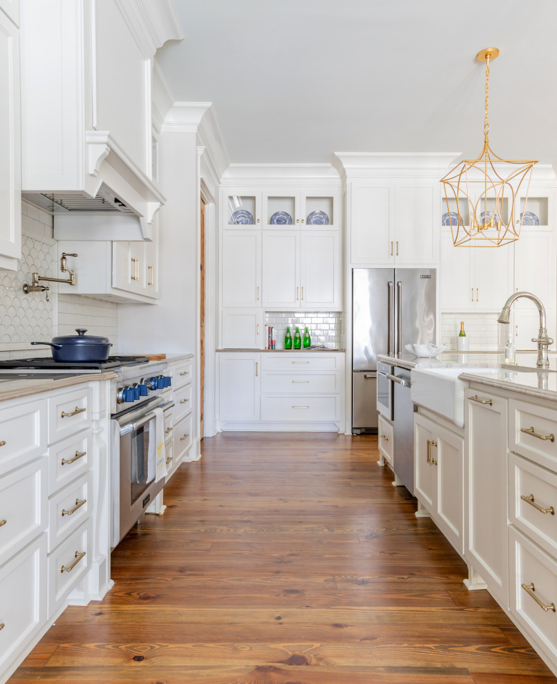 Large traditional l-shaped open plan kitchen in Atlanta with a belfast sink, shaker cabinets, white cabinets, engineered stone countertops, white splashback, porcelain splashback, stainless steel appliances, medium hardwood flooring, an island, brown floors and beige worktops.