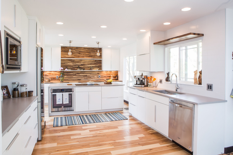 Photo of a medium sized contemporary u-shaped kitchen/diner in Denver with a submerged sink, flat-panel cabinets, white cabinets, engineered stone countertops, white splashback, ceramic splashback, black appliances, light hardwood flooring, an island, brown floors and grey worktops.