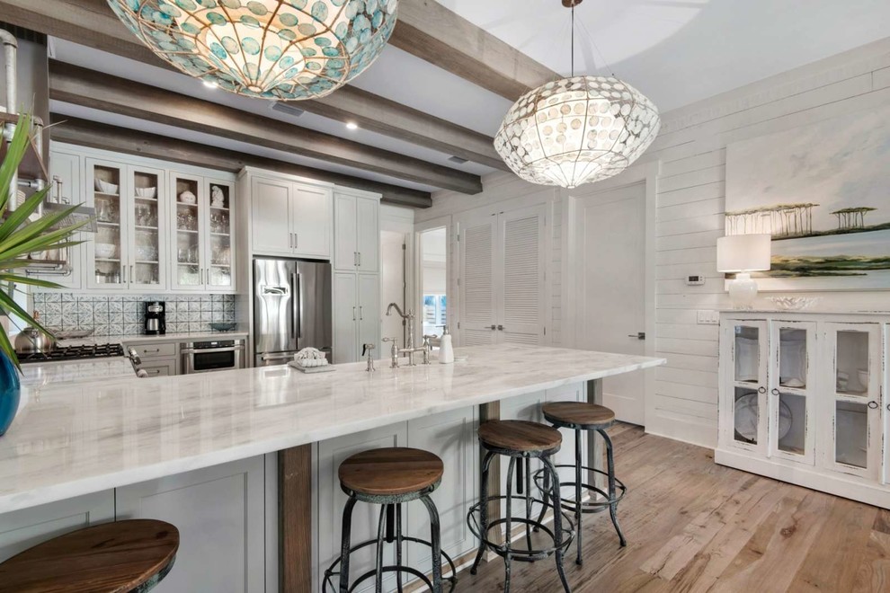 Example of a mid-sized beach style u-shaped light wood floor and brown floor enclosed kitchen design in Miami with a farmhouse sink, shaker cabinets, white cabinets, quartzite countertops, white backsplash, wood backsplash, stainless steel appliances, a peninsula and white countertops
