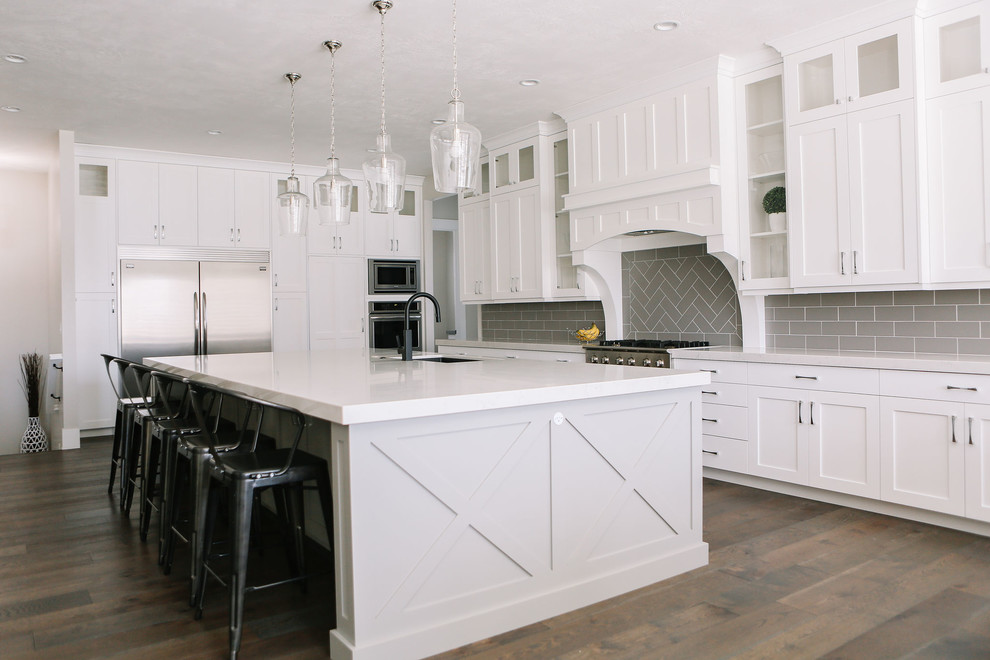 Exempel på ett stort klassiskt vit vitt kök, med en undermonterad diskho, luckor med infälld panel, vita skåp, bänkskiva i kvartsit, grått stänkskydd, stänkskydd i tunnelbanekakel, rostfria vitvaror, mörkt trägolv, en köksö och brunt golv