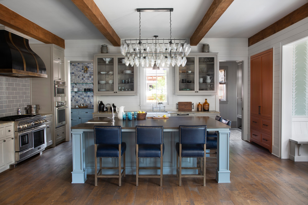 Inspiration for a large timeless dark wood floor and brown floor kitchen remodel in Denver with a farmhouse sink, solid surface countertops, stainless steel appliances, an island, brown countertops, glass-front cabinets, gray cabinets, gray backsplash and subway tile backsplash