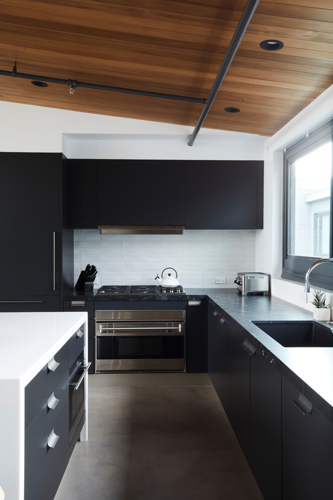 На фото: угловая кухня в современном стиле с врезной мойкой, плоскими фасадами, белым фартуком, техникой из нержавеющей стали, бетонным полом, островом, серым полом и черно-белыми фасадами