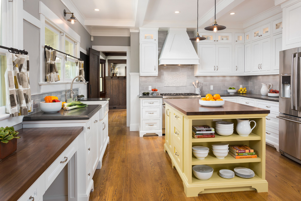 Example of a mid-sized classic u-shaped medium tone wood floor eat-in kitchen design in Los Angeles with a farmhouse sink, recessed-panel cabinets, white cabinets, granite countertops, gray backsplash, ceramic backsplash, stainless steel appliances and an island