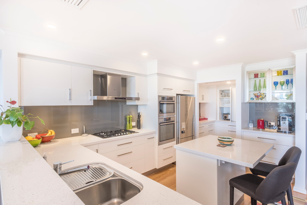 This is an example of a contemporary kitchen in Perth with a submerged sink, flat-panel cabinets, white cabinets, engineered stone countertops, grey splashback, glass sheet splashback, stainless steel appliances, an island and white worktops.