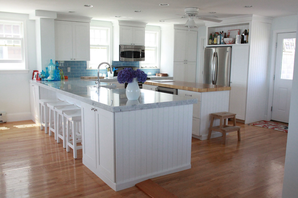 Example of a large beach style u-shaped light wood floor and brown floor eat-in kitchen design in Boston with a farmhouse sink, shaker cabinets, white cabinets, quartz countertops, blue backsplash, subway tile backsplash, stainless steel appliances and an island