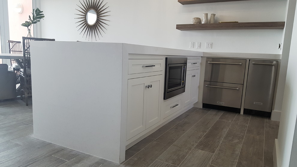 Photo of a large coastal u-shaped kitchen/diner in Miami with a belfast sink, shaker cabinets, white cabinets, quartz worktops, white splashback, metro tiled splashback, stainless steel appliances, ceramic flooring, an island and grey floors.