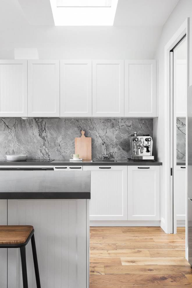 Photo of a large contemporary galley kitchen pantry in Melbourne with a submerged sink, shaker cabinets, white cabinets, engineered stone countertops, grey splashback, stone slab splashback, stainless steel appliances, light hardwood flooring, an island and grey worktops.