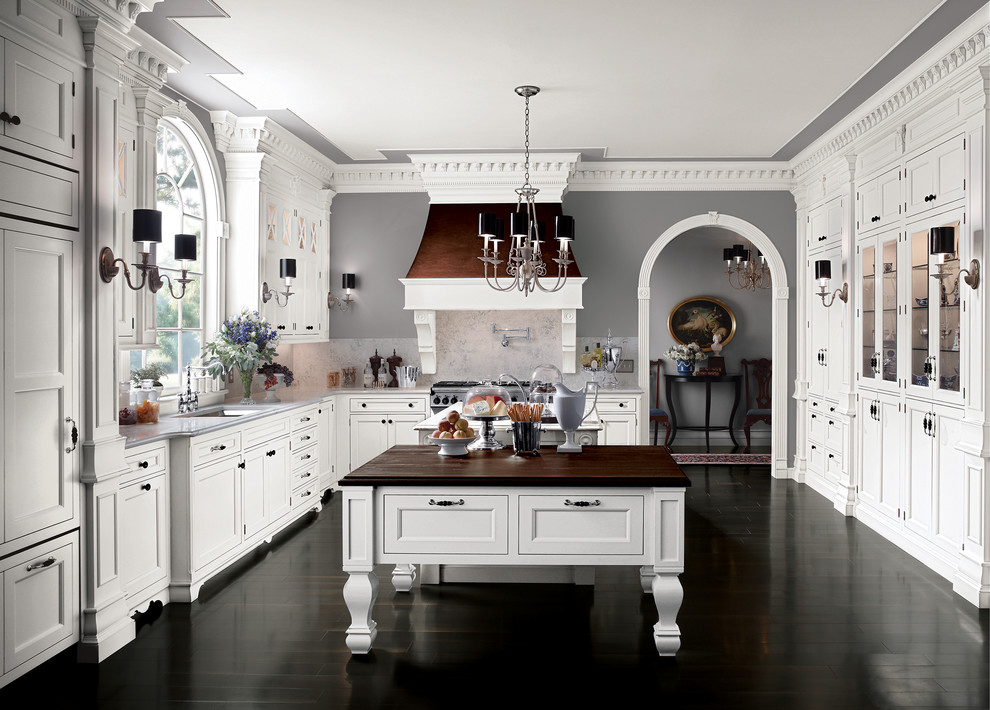 На фото: п-образная кухня в классическом стиле с врезной мойкой, фасадами с утопленной филенкой, белыми фасадами, деревянной столешницей, серым фартуком, техникой под мебельный фасад и мойкой у окна с