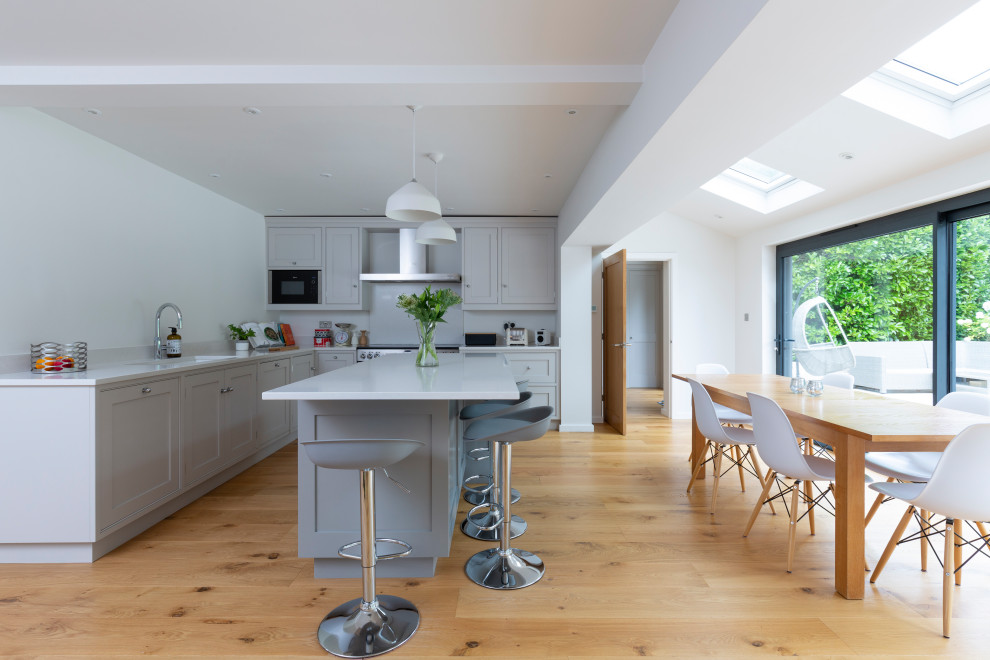 Photo of a large traditional l-shaped kitchen/diner with a submerged sink, shaker cabinets, grey cabinets, white splashback, stainless steel appliances, medium hardwood flooring, an island, brown floors and white worktops.