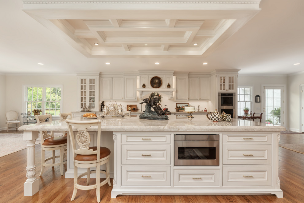 Klassisk inredning av ett mycket stort vit vitt kök, med luckor med upphöjd panel, vita skåp, bänkskiva i kvartsit, vitt stänkskydd, mellanmörkt trägolv, en köksö och brunt golv
