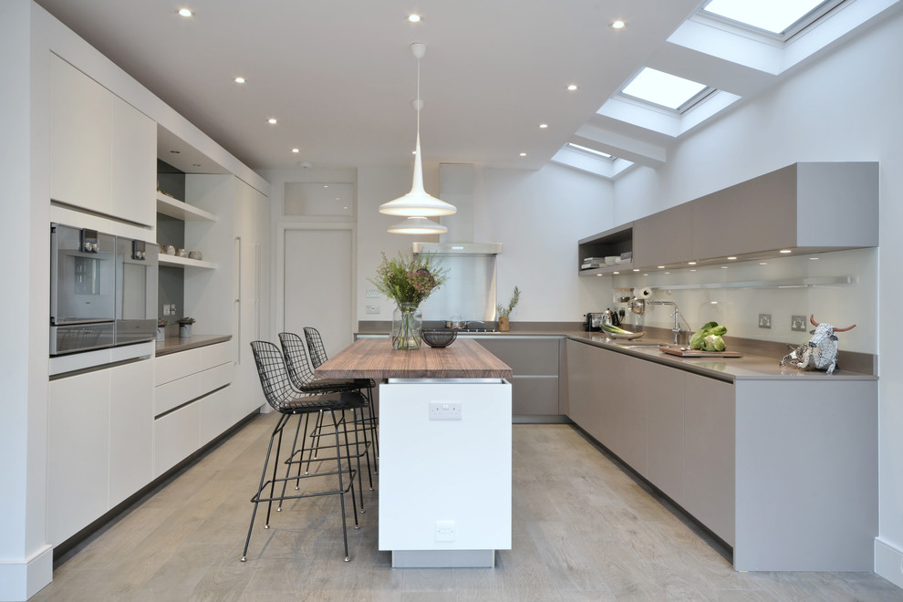 Example of a trendy u-shaped light wood floor kitchen design in Gloucestershire with flat-panel cabinets, gray cabinets, gray backsplash, stainless steel appliances and an island