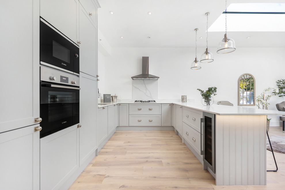 This is an example of a large classic grey and white u-shaped open plan kitchen in London with a submerged sink, shaker cabinets, grey cabinets, grey splashback, stainless steel appliances, light hardwood flooring, a breakfast bar, beige floors and white worktops.