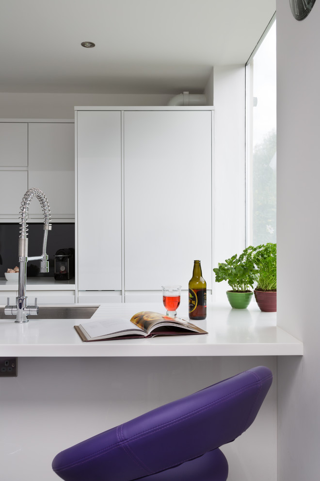 Trendy galley eat-in kitchen photo in London with an integrated sink, flat-panel cabinets, white cabinets, solid surface countertops, gray backsplash and stainless steel appliances