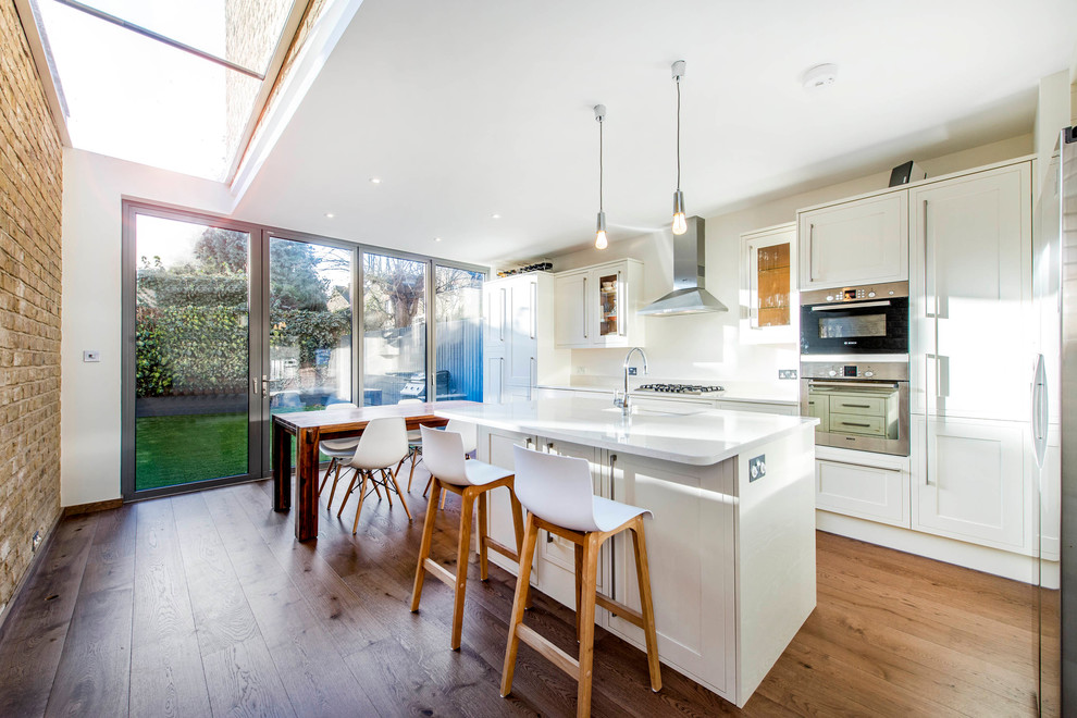 Zweizeilige Klassische Wohnküche mit Schrankfronten im Shaker-Stil, weißen Schränken, Küchengeräten aus Edelstahl, braunem Holzboden und Kücheninsel in London