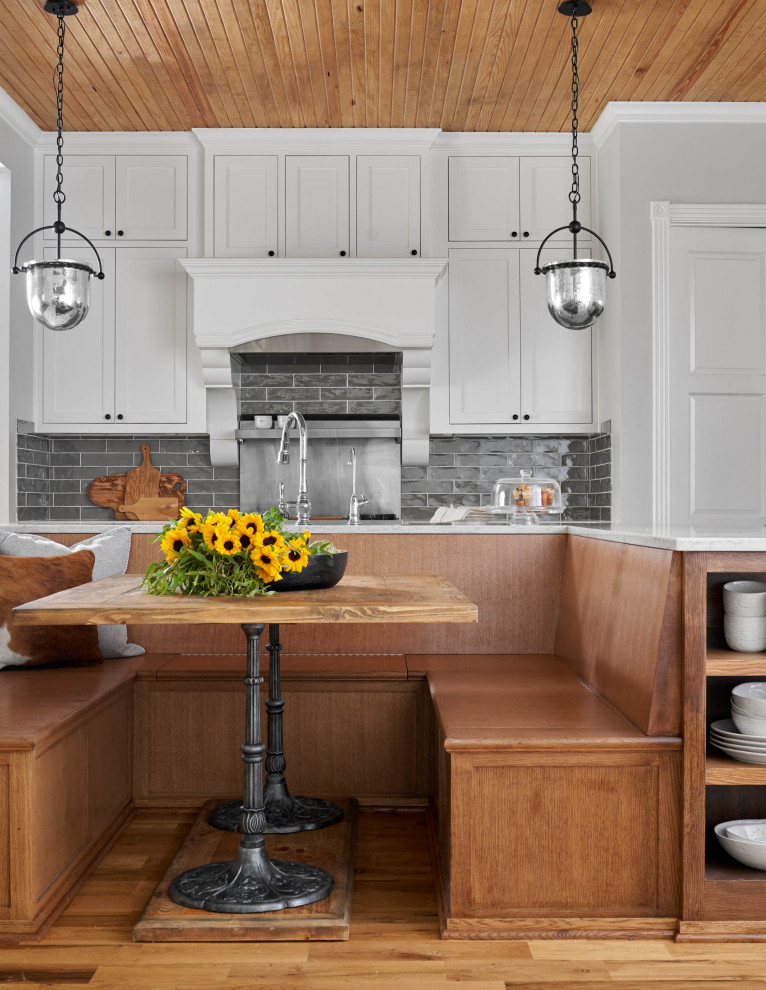 This is an example of a medium sized rural u-shaped kitchen/diner in Dallas with a belfast sink, shaker cabinets, white cabinets, engineered stone countertops, grey splashback, metro tiled splashback, stainless steel appliances, medium hardwood flooring, an island, brown floors and white worktops.