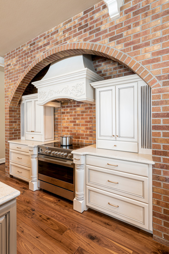 Cette image montre une cuisine traditionnelle avec un placard avec porte à panneau encastré, un plan de travail en quartz modifié, une crédence en brique, un électroménager en acier inoxydable, un sol en bois brun et un plan de travail blanc.