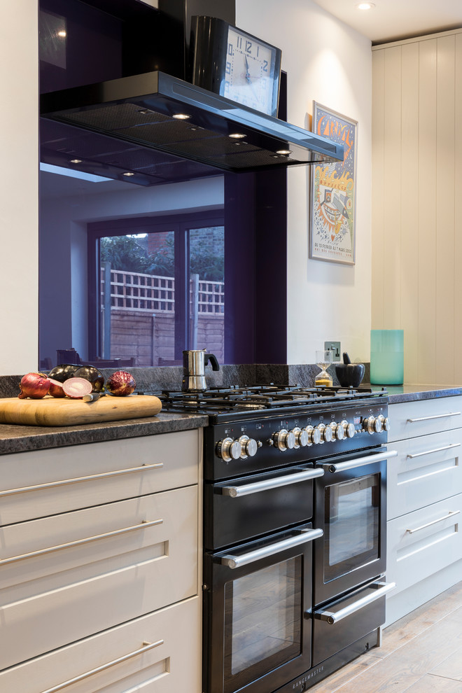 This is an example of a large contemporary galley kitchen/diner in London with an integrated sink, shaker cabinets, grey cabinets, black appliances, medium hardwood flooring, an island and brown floors.