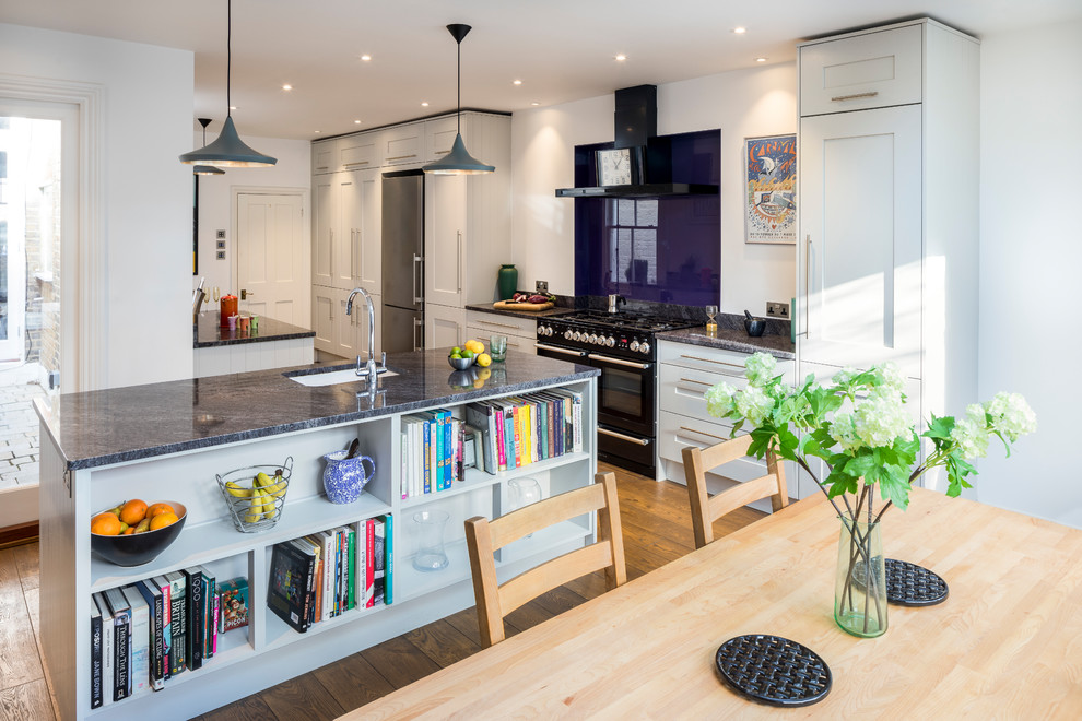 This is an example of a classic kitchen/diner in London with a submerged sink, shaker cabinets, grey cabinets, glass sheet splashback, black appliances, medium hardwood flooring, an island, brown floors and grey worktops.