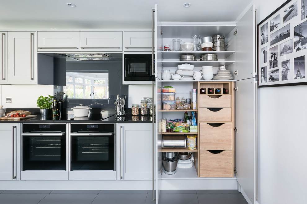 Klassische Küche mit Schrankfronten im Shaker-Stil, grauen Schränken, Küchenrückwand in Grau, Glasrückwand, schwarzen Elektrogeräten und grauem Boden in London