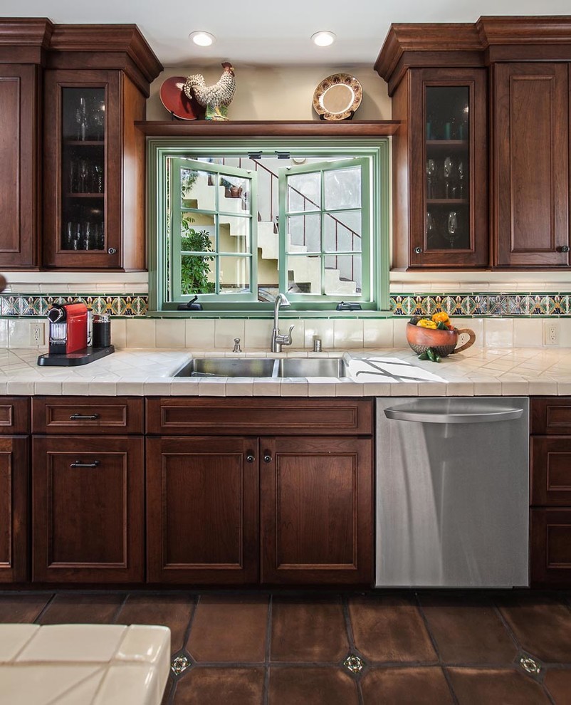 Photo of a medium sized mediterranean galley enclosed kitchen in Los Angeles with a double-bowl sink, recessed-panel cabinets, dark wood cabinets, tile countertops, green splashback, ceramic splashback, stainless steel appliances and ceramic flooring.