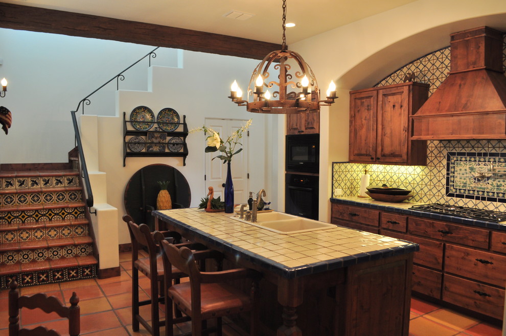 オースティンにある高級な中くらいなサンタフェスタイルのおしゃれなキッチン (中間色木目調キャビネット、タイルカウンター、テラコッタタイルの床、ダブルシンク、レイズドパネル扉のキャビネット、マルチカラーのキッチンパネル、セラミックタイルのキッチンパネル、黒い調理設備) の写真