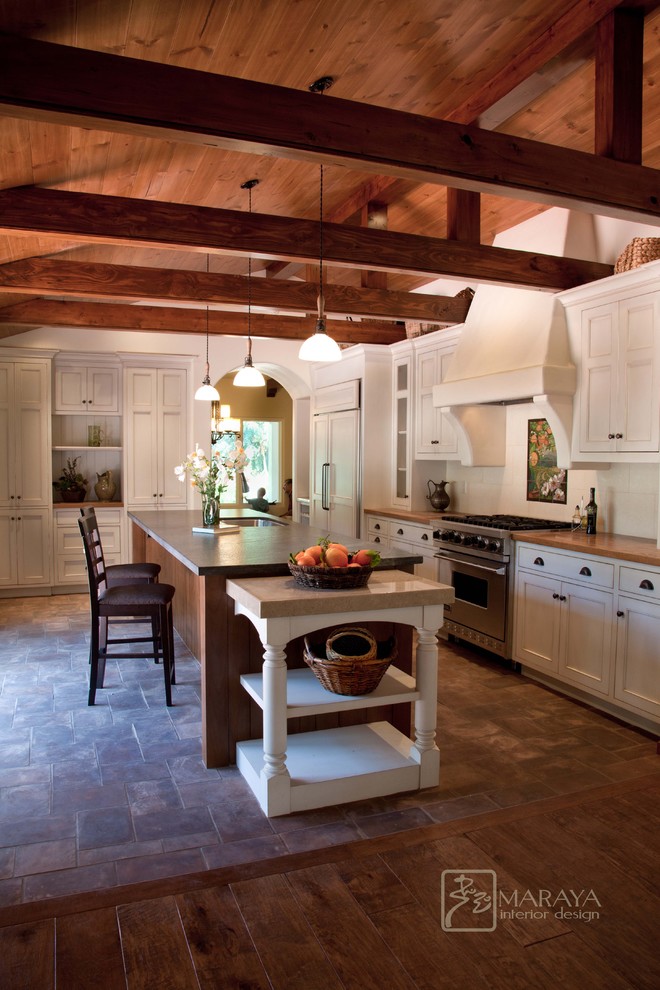 Example of a mid-sized country l-shaped ceramic tile open concept kitchen design in Santa Barbara with a single-bowl sink, recessed-panel cabinets, white cabinets, wood countertops, beige backsplash, stone tile backsplash, stainless steel appliances and an island