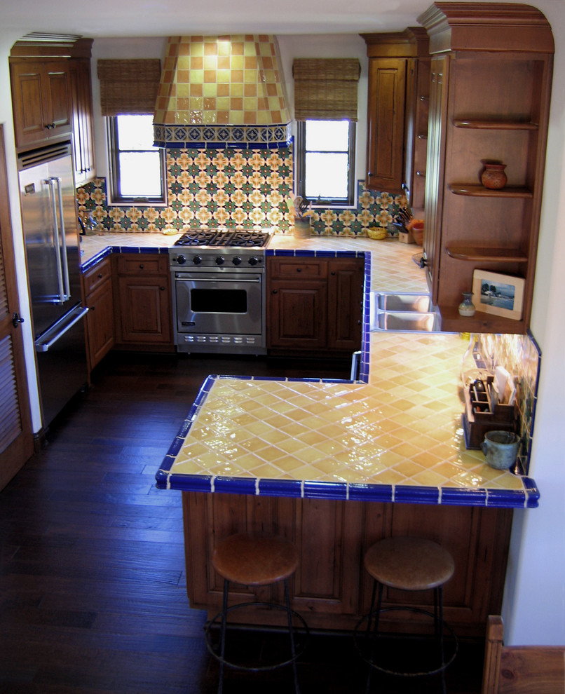 Inredning av ett medelhavsstil litet gul gult kök, med en dubbel diskho, luckor med upphöjd panel, skåp i mellenmörkt trä, kaklad bänkskiva, stänkskydd i keramik, rostfria vitvaror, mörkt trägolv, en halv köksö, flerfärgad stänkskydd och brunt golv
