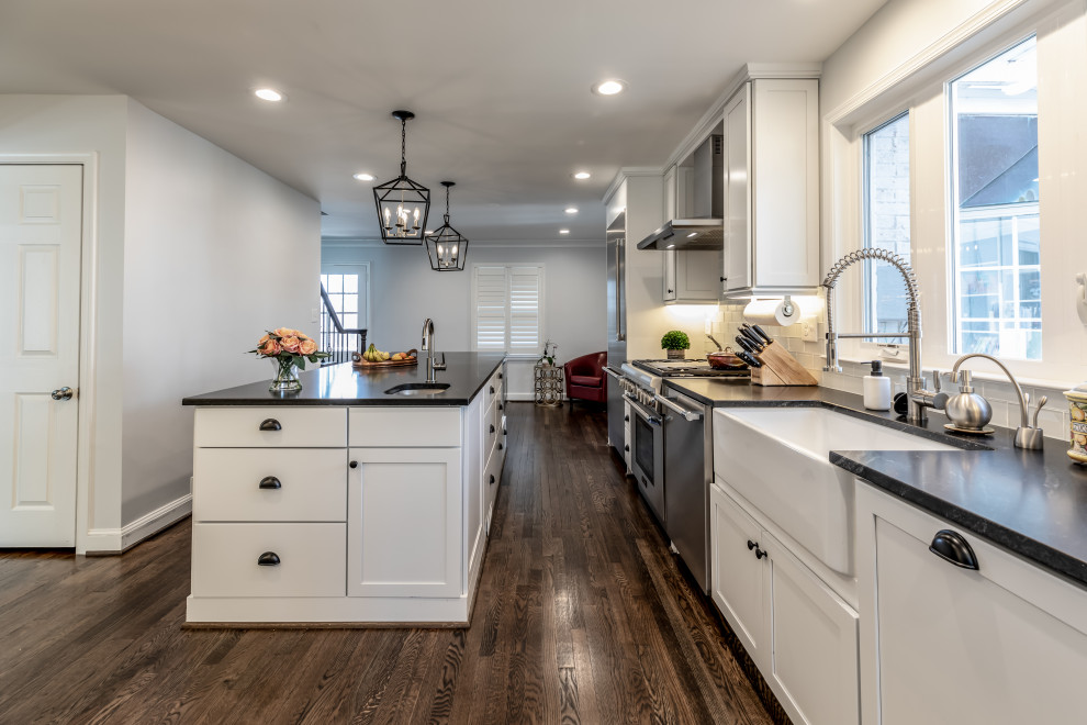 Example of a large transitional galley medium tone wood floor and brown floor kitchen design in DC Metro with a farmhouse sink, shaker cabinets, white cabinets, gray backsplash, subway tile backsplash, stainless steel appliances, an island and black countertops