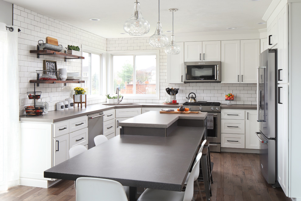 Bild på ett lantligt grå grått kök, med en enkel diskho, skåp i shakerstil, vita skåp, vitt stänkskydd, stänkskydd i tunnelbanekakel, rostfria vitvaror, mörkt trägolv, en köksö och brunt golv